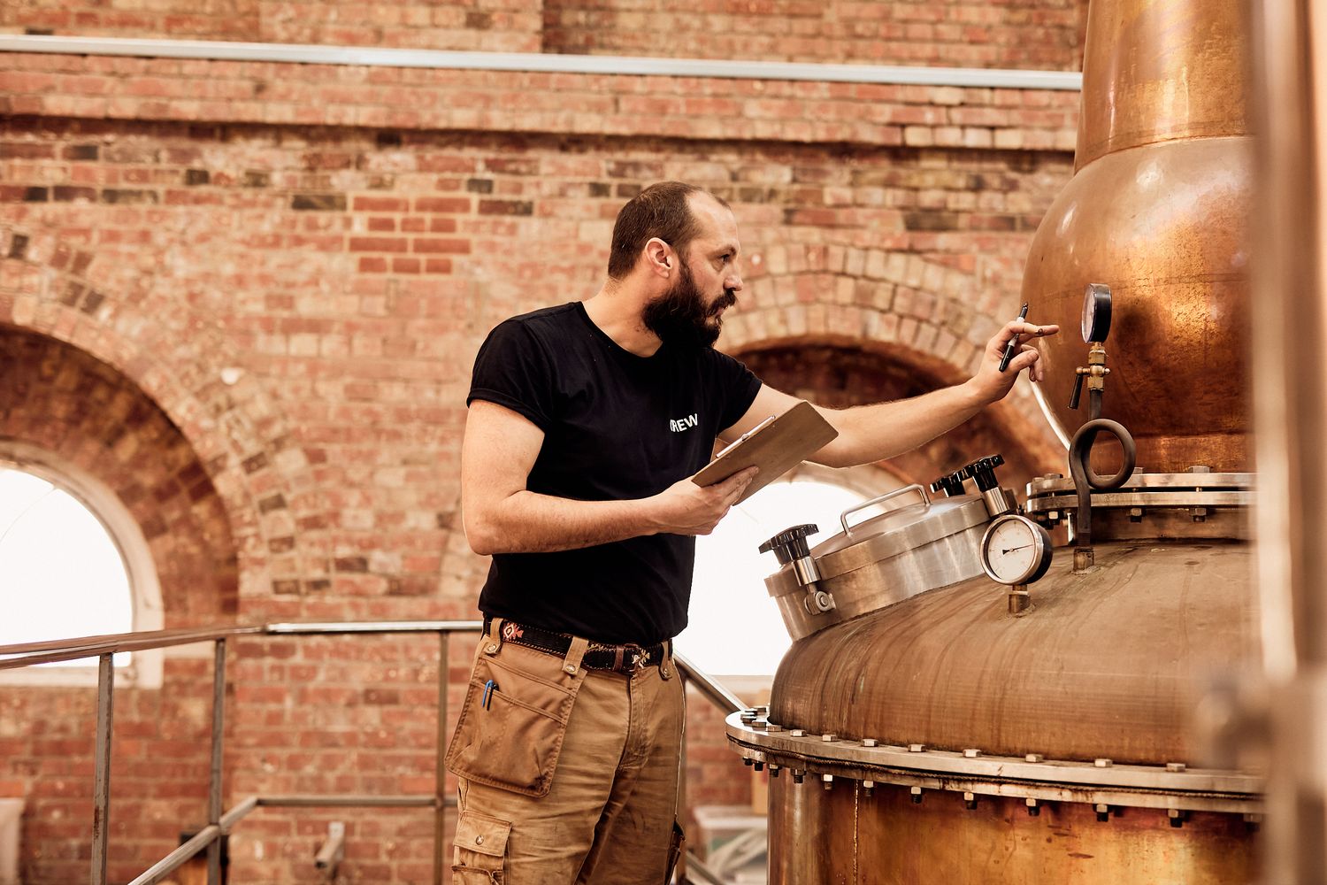 A man in a black t-shirt and beige pants is standing next to a large copper tank in a rustic setting. He is holding a clipboard and appears to be examining the tank. The tank is large and metallic, with a round dial on top. The man is standing in front of a brick wall, which has a window. The image captures a moment of concentration and attention to detail in a setting that suggests a distillery or brewery. [G]