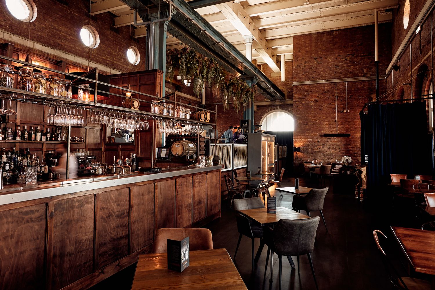 A cozy restaurant with rustic charm, featuring a wooden bar, tables, and chairs, adorned with plants and hanging wine glasses. [G]