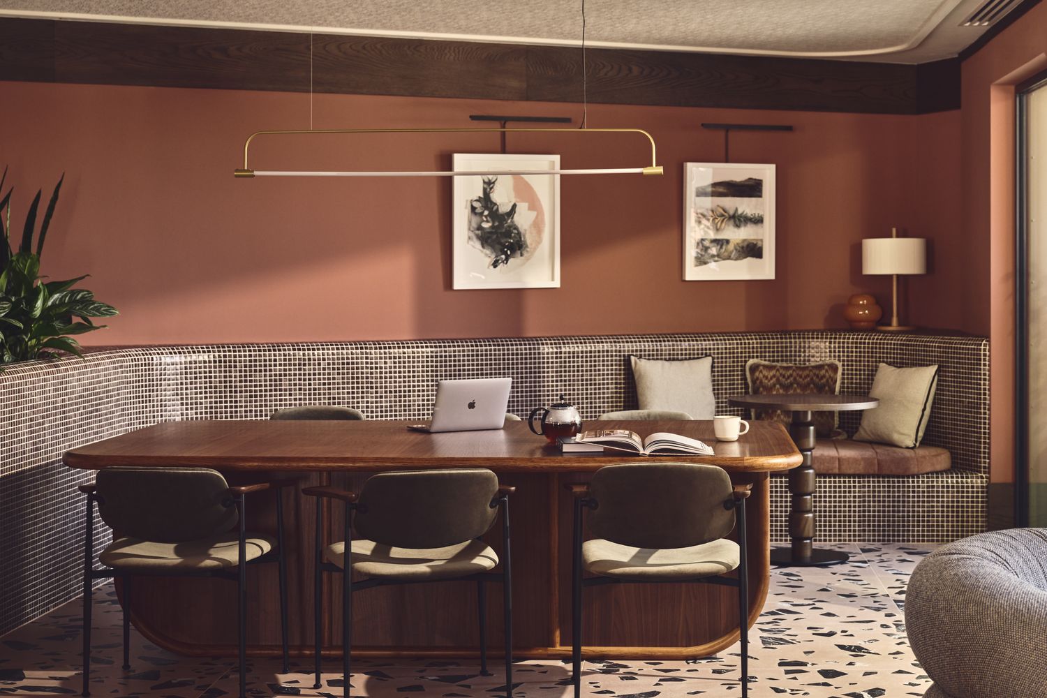 A modern living room with a large wooden table, four chairs, and a laptop on the table. The wall has artwork and a lamp. [G]