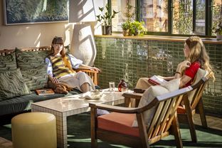 The image features two women lounging in a cozy living room. One woman is wearing a striped vest and a white skirt, while the other is in a yellow and white striped vest and a white skirt. They are surrounded by a green and white tiled wall, a wooden coffee table, and a vase with a plant. The room is illuminated by natural light streaming in through a window. [G]