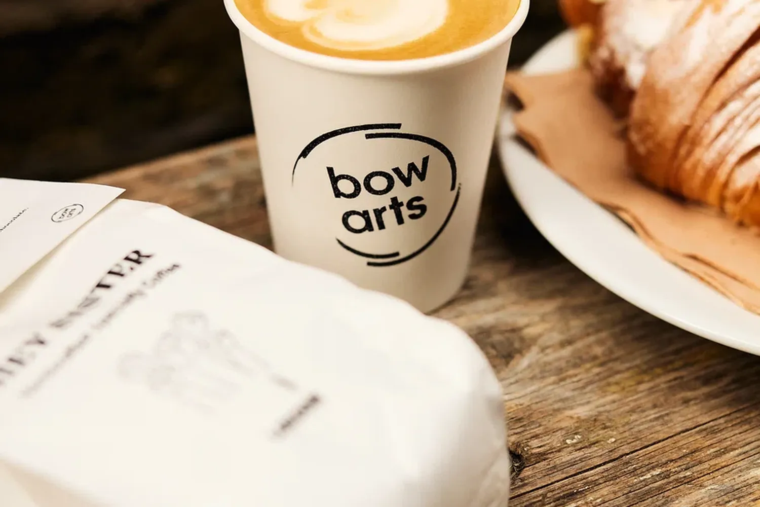 A coffee cup with a "Wol-deptford" logo, a croissant on a plate, and a white napkin with a credit card. [G]