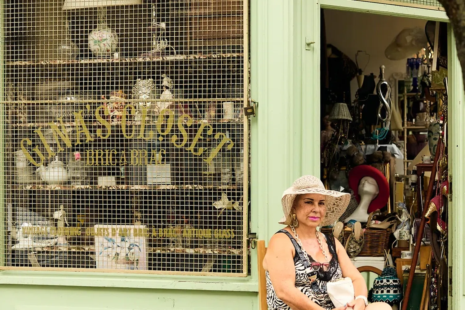 A woman sits on a chair outside a shop named "Wol-deptford" with a variety of hats and accessories displayed in the window. [G]