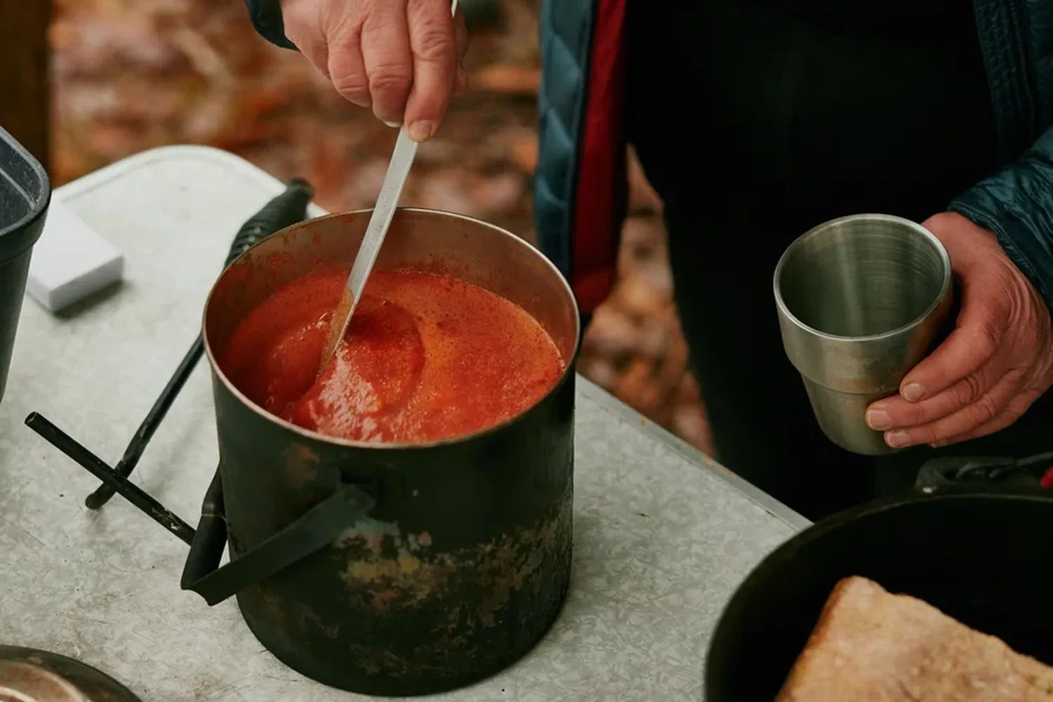 A person is cooking in a wooden outdoor kitchen, using a spoon to stir a red sauce in a pot on a stove. They are holding a silver cup and a black bowl. There are two pots on the stove, one black and one red. The person is wearing a blue jacket and a red scarf. The background shows trees and leaves. [G]