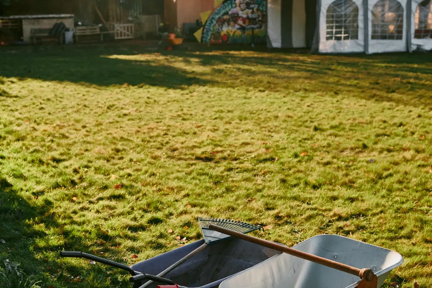 A sunny day in a lush green yard with a white tent in the background. A wheelbarrow and shovel are placed on the grass, ready for use. [G]