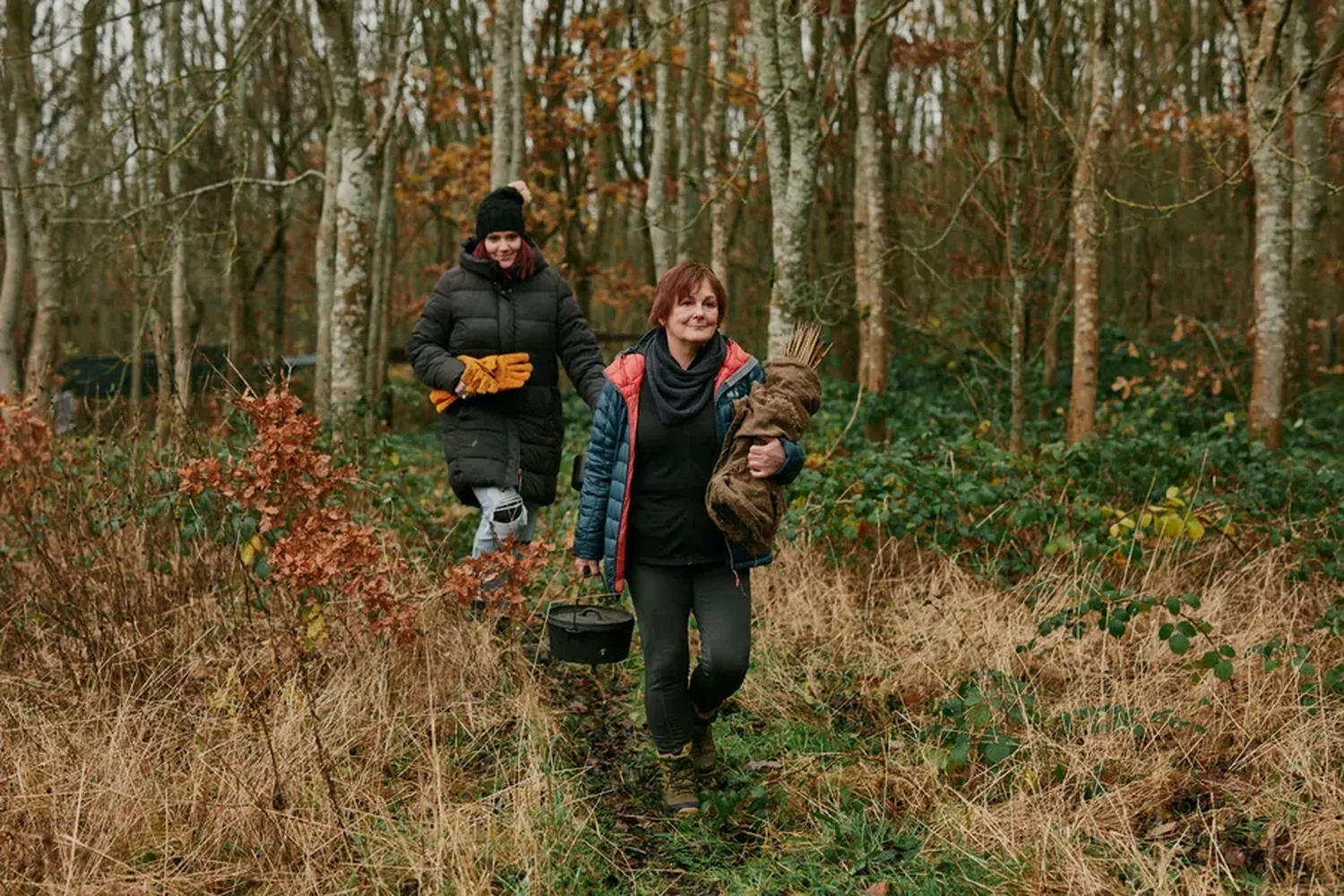 A woman in a black jacket and a man in a black hat and jacket walk through a forest, carrying a brown stuffed animal and a black bucket. [G]