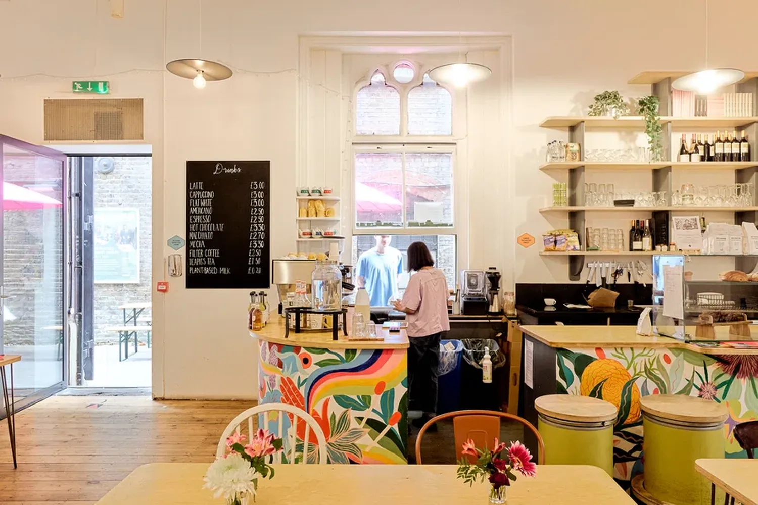 A woman in a pink shirt is working behind a colorful counter at a coffee shop. The counter has a menu board and a coffee machine. There are flowers on the counter and a window with a view of the outside. The shop has a chalkboard menu and a blackboard menu. The woman is standing behind the counter. [G]