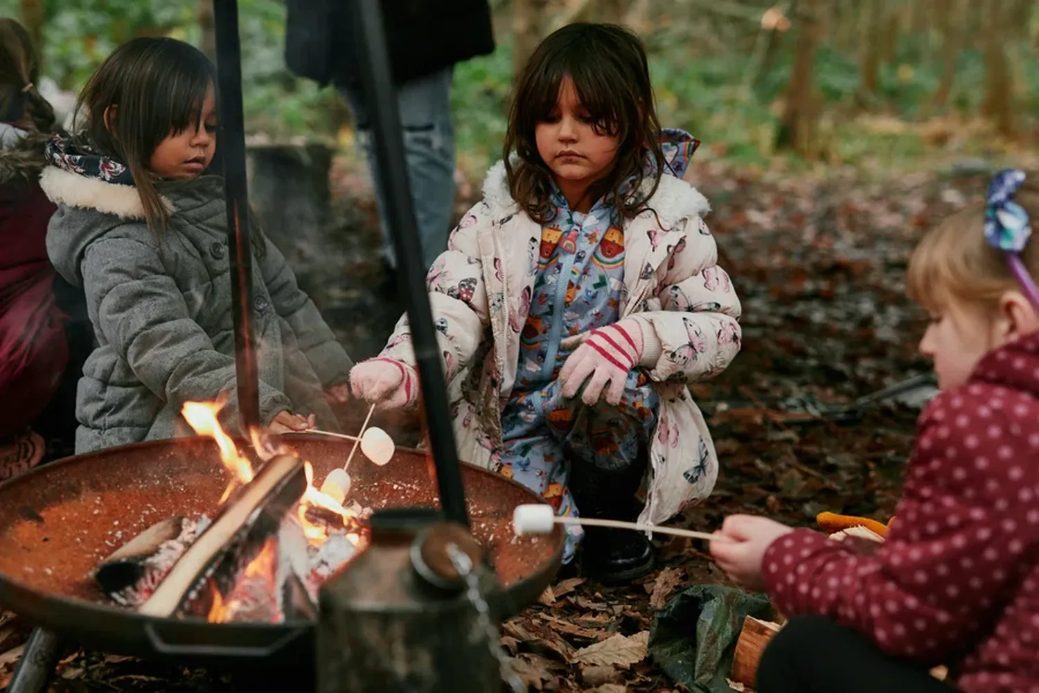 A young girl in a white coat and pink scarf is cooking marshmallows on a stick over a campfire in a wooded area. [G]