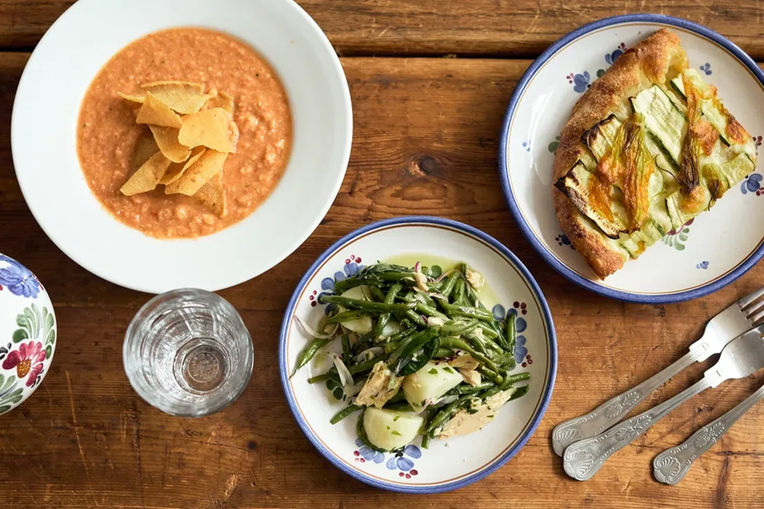 A rustic wooden table is set with a variety of dishes. There's a bowl of soup with a crusty crust, garnished with green beans and a fried egg. Next to it, there's a plate of roasted potatoes and a side of green beans. A glass of water is also present on the table. [G]