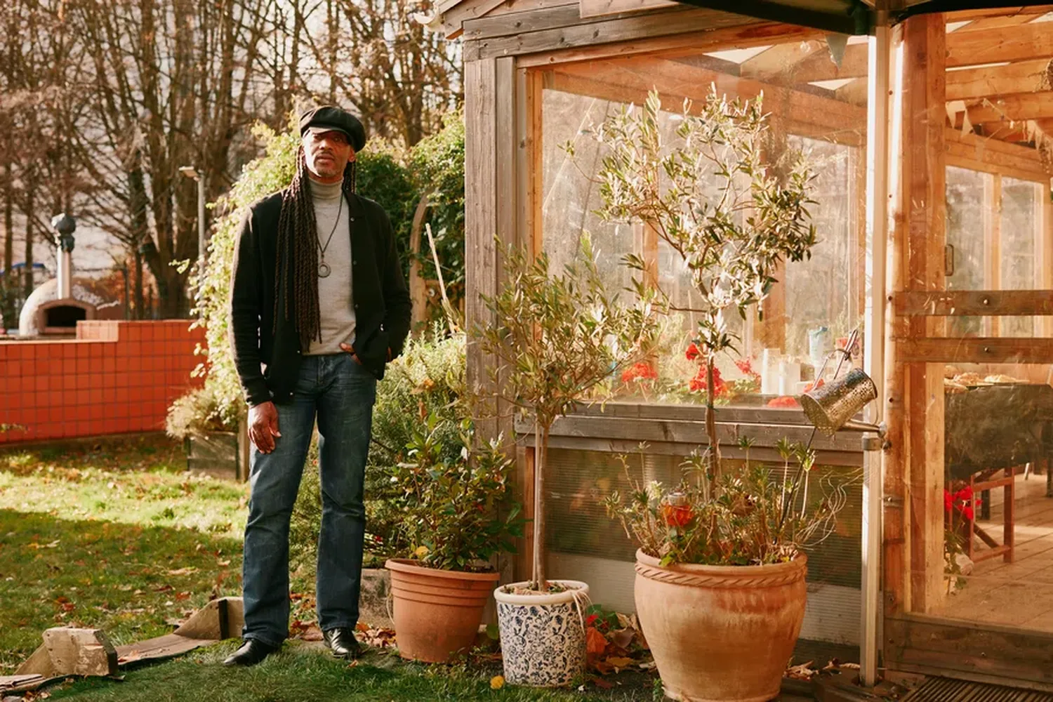 A man with dreadlocks, wearing a black hat, a white shirt, blue jeans, and a black jacket, stands in front of a wooden greenhouse with a glass window. He is surrounded by potted plants and a red flower. [G]