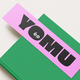 A green book with a pink bookmark showing the word "YOMU" in bold black letters, featuring a cute face in the letter "O".