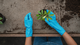 Person wearing blue gloves tending to a small potted plant on a table, surrounded by soil.