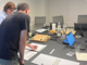Two people play a strategy board game in an office setting, surrounded by laptops, snacks, and drinks on a conference table.