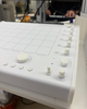White music controller with multiple knobs, buttons, and a grid interface on a desk. Background shows blurred office setting.