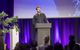 A keynote speaker presenting on stage at an S•HUB event, surrounded by greenery and vibrant purple lighting, symbolizing sustainability and innovation.