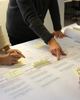 Three people collaborate on a large paper with diagrams and sticky notes, taking notes and discussing plans in an office setting.