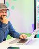 Smiling man in a cap and denim shirt sitting at a table with a laptop, a phone nearby, colorful background, and blurred figure in doorway.