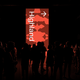 Silhouetted crowd standing in front of a tall, illuminated red sign displaying the word "Highland" with an upward arrow.