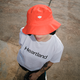 Person in a white "Heartland" shirt wearing a bright red bucket hat with a heart logo, standing on a textured pavement.