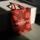 Red tote bag with abstract design and the word "Heartland" printed on it, leaning against a white geometric structure in soft lighting.