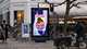 Street scene with pedestrians, cyclists, and a digital billboard displaying an advertisement for a book titled "Maame" by Jessica George.