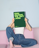 Person sitting on a pink sofa, holding a green sign covering their face. The sign reads "DIN GUIDE TIL DIT JOB" in bold letters.