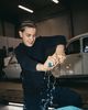 Person squeezing a wet cloth over a blue bucket, with water splashing, in a garage with a vintage car in the background.