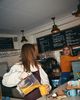 A woman spills orange smoothie on a counter while pouring from a blender, as another woman in a café smiles in the background.
