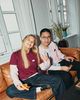 Two people sitting on a brown couch, smiling and eating pizza. One person holds a peace sign. Bright, cozy room with windows and a plant.