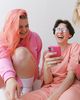 Two people laughing, wearing pink outfits. One holds a pink phone. The image has a hexagonal frame with a purple background.
