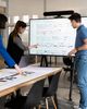 Three people discuss a project, pointing at diagrams on a digital display and papers on a table in a modern office setting.