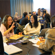 A group of three engage in a lively discussion at a workshop table cluttered with laptops, papers, and cards, conveying collaboration and focus.