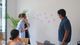 People in a meeting room discussing ideas with sticky notes on the wall. Laptops are on the table, and a plant is in the background.
