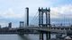The Manhattan Bridge spans over the East River, with a city skyline in the background under a partly cloudy sky.