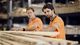 Two workers in orange safety vests inspect wooden planks in a warehouse, focusing on quality and alignment.