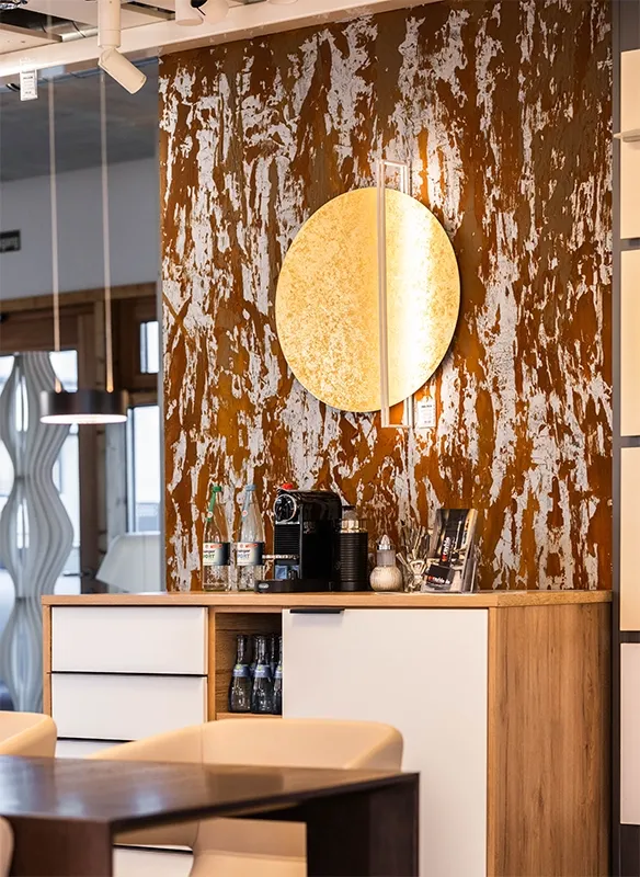 Lichthaus Melzer Ausstellung mit modernem Interieur mit einem Holzschrank, einer Kaffeemaschine und einer runden Wandleuchte an einer strukturierten, rostfarbenen Wand. Beige Stühle sind teilweise sichtbar.