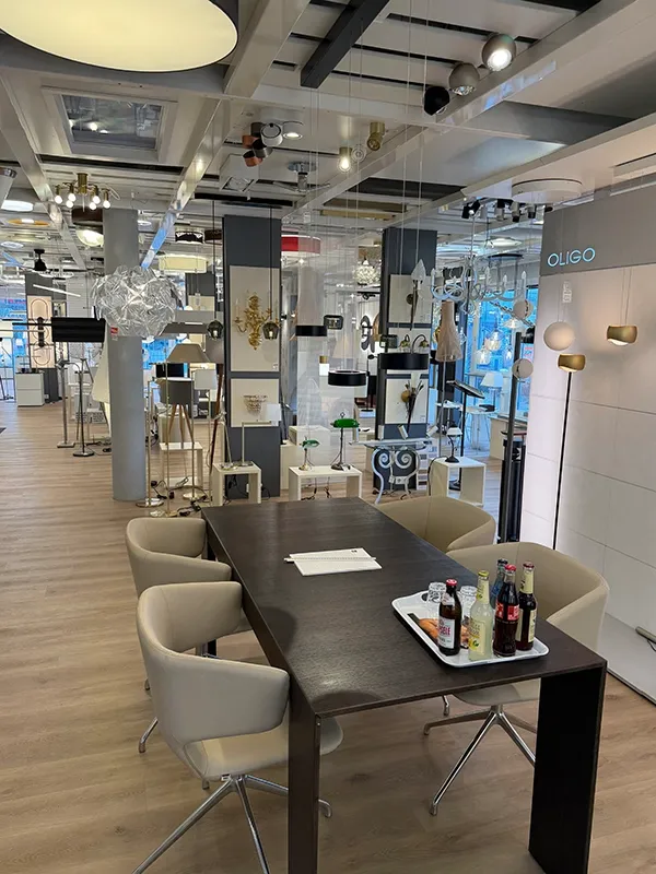 Lichthaus Melzer Ausstellung -2- Ein moderner Beleuchtungs-Showroom mit verschiedenen Leuchten, einem dunklen Holztisch mit Getränken, umgeben von beigen Stühlen und beleuchteten Deckenpaneelen.