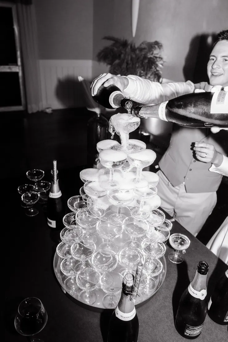 Personne versant du champagne dans une pyramide de verres lors d'un événement, avec des bouteilles sur la table, dans un décor noir et blanc.