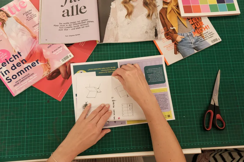 auf einer dunkelgrünen Schneidematte sieht man zwei Hände, die ein Blatt Papier, daneben einige Magazine und eine Schere