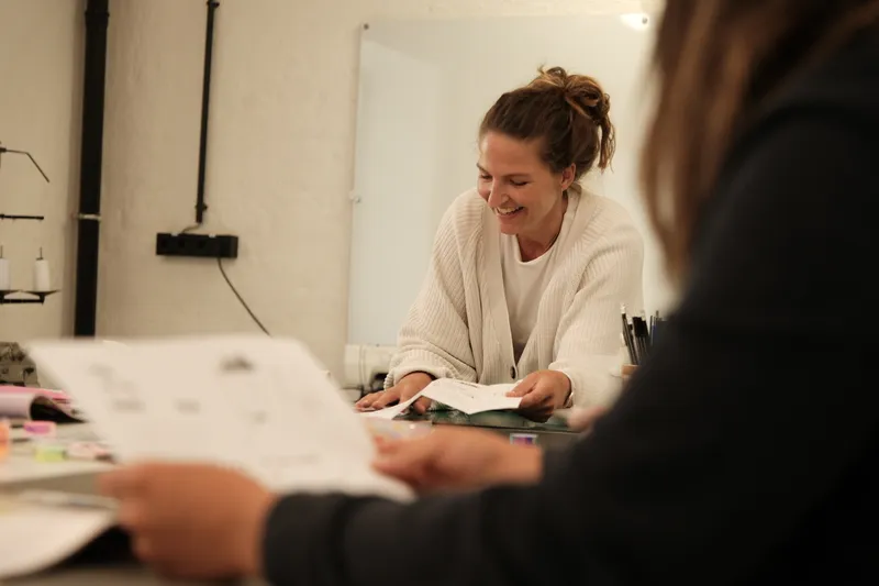 Workshopsituation, unscharf im Vordergrund eine Teilnehmer:in mit Unterlagen in der Hand, im Fokus steht eine gut gelaunte Workshopleiterin, die durch ihre Unterlagen blättert