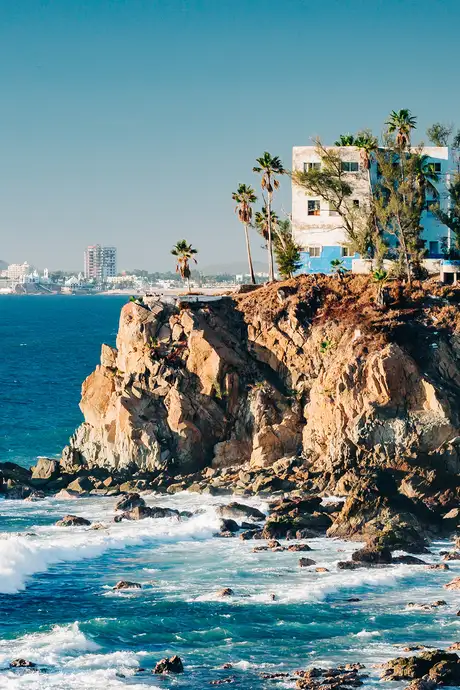 Cliffside building with palm trees overlooking a rocky shoreline and blue ocean waves under a clear sky.
