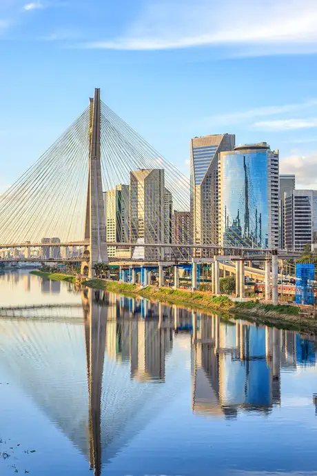 Cable-stayed bridge over a calm river reflecting modern skyscrapers and a blue sky with clouds.