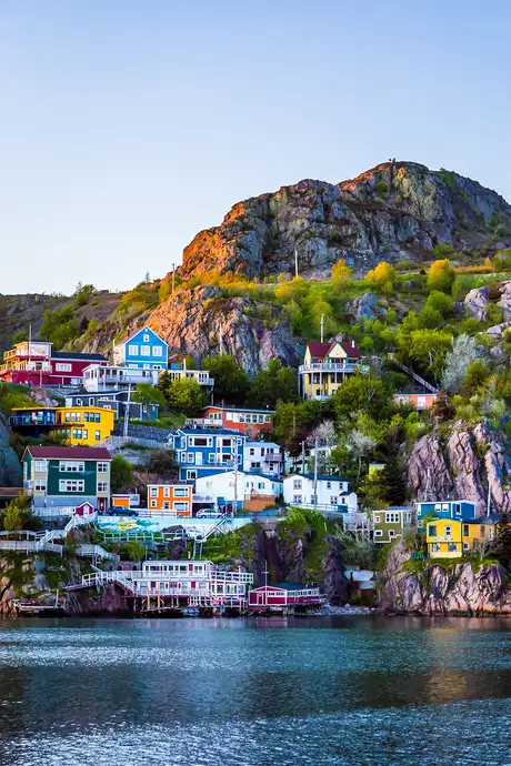 Colorful houses on a rocky hillside overlook calm water, with a clear blue sky above. Lush greenery is interspersed among the buildings.
