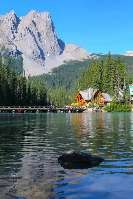 A serene mountain lake with a wooden cabin, surrounded by pine trees, under a clear blue sky and rocky mountains in the background.