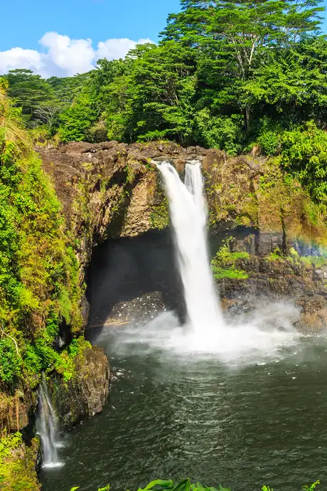 A waterfall cascades into a pool surrounded by lush green foliage and rocky cliffs under a blue sky.