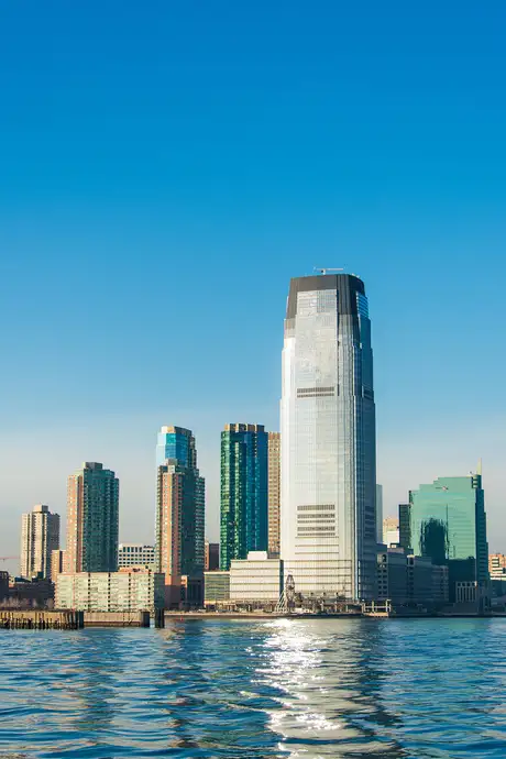 Skyline of modern skyscrapers reflecting on calm water under a clear blue sky.