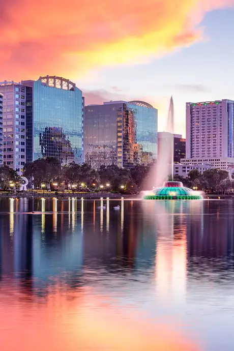 City skyline at sunset with colorful clouds, reflecting on a calm lake. A fountain is illuminated in the center, surrounded by modern buildings.