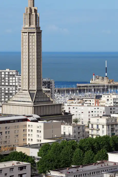 Aerial view of a coastal city with a tall, modern church tower, residential buildings, and a marina by the sea under a clear blue sky.