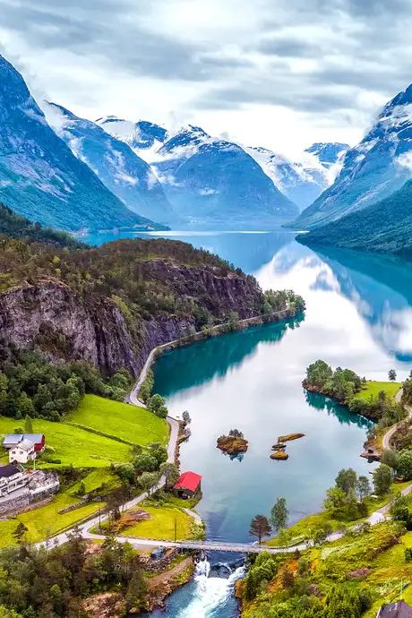 Scenic view of a winding river through lush green valleys, surrounded by snow-capped mountains under a cloudy sky.