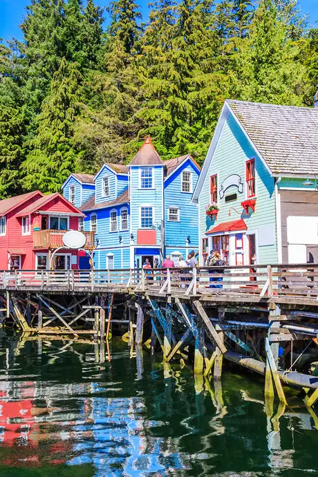 Colorful wooden buildings on stilts line a waterfront, surrounded by lush trees and reflected in the calm water below.