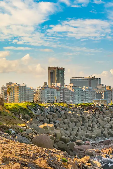 Rocky shoreline with a view of a cityscape in the background, featuring modern buildings under a blue sky with scattered clouds.